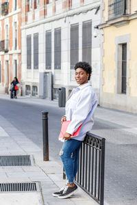 Positive african american female student with red folder in hand looking at camera with smile while standing on street of city