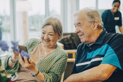 Smiling senior woman sharing smart phone with man while sitting at retirement home