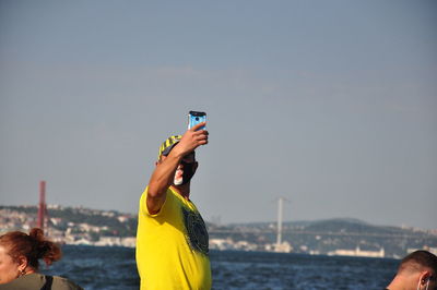 Man photographing with mobile phone by sea against sky