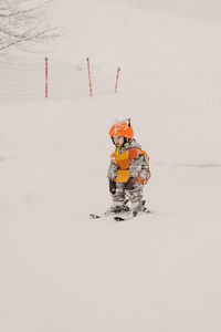 Full body of adorable little child in warm winter overalls and protective helmet standing on skis while practicing skiing on snowy terrain