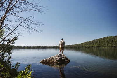Rear view of hiker standing on rock amidst lake against clear sky