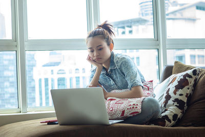 Young woman using phone while sitting on window