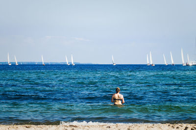 Rear view of woman in sea looking at sailboats against sky