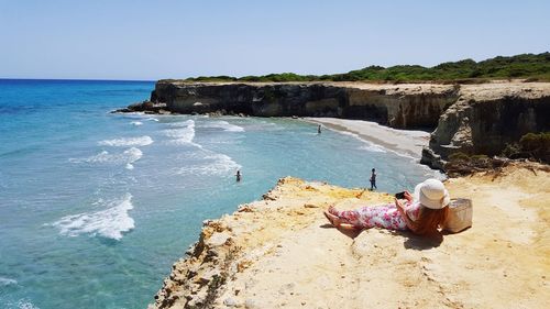 Woman photographing through phone while lying on cliff by sea against clear sky