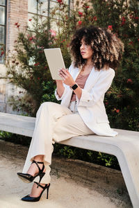 Full body hispanic businesswoman in white suit with curly hair sitting on bench and browsing tablet on street