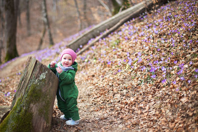 Cute toddler baby wearing green overall and pink hat in spring forest full of wild irises.