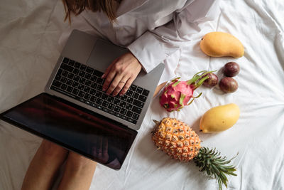 Midsection of woman using laptop sitting by fruits on bed