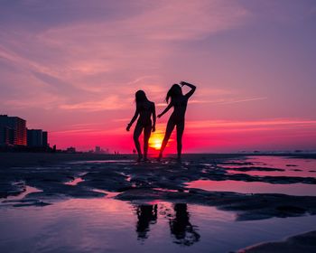 Silhouette females standing at beach against sky during sunset