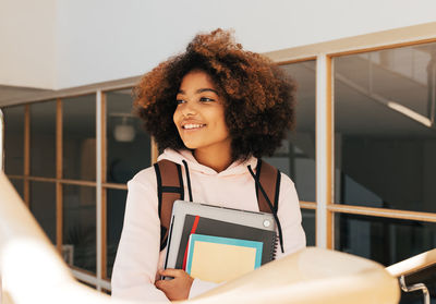 Happy teenage girl with laptop and books standing against glass wall