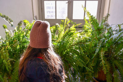 Rear view of woman wearing knit hat standing by potted plants against window