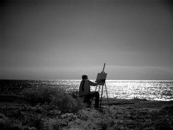 Man painting on canvas while sitting on field by sea against sky