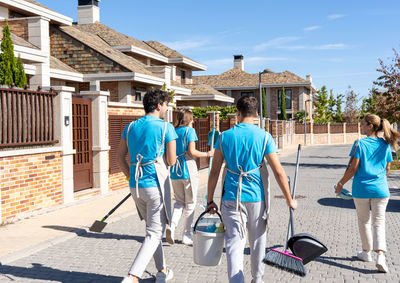 Back view of unrecognizable young cleaning crew in uniform carrying brooms with scoops and buckets while walking on paved street near residential houses on sunny day