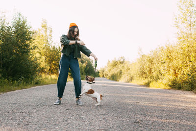 A beautiful teenage girl in a green jacket and orange hat plays with her dog while walking 