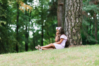 A child girl leans her back against a tree and sits on the grass in summer in an urban landscape