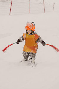 Full body of concentrated little child in warm clothes and protective orange helmet and vest holding brushes while practicing skiing on snowy slope on winter day