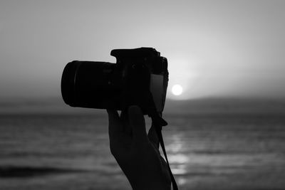 Cropped hand of woman holding dslr camera against sky during sunset