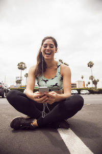 Young woman laughing while using smart phone sitting on street