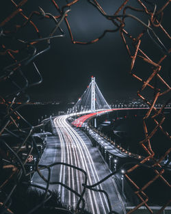 View of light trails on bridge seen through chainlink fence at night