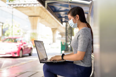 Side view of woman wearing mask using laptop outdoors
