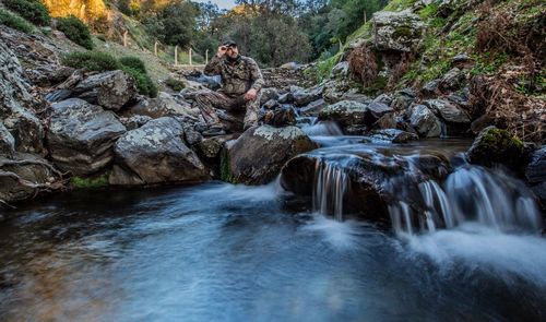 Full length of thoughtful man sitting on rock by stream in forest