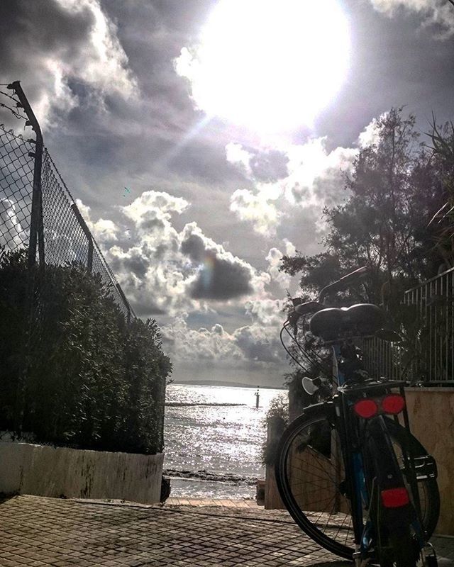 sky, water, cloud - sky, railing, cloudy, sunlight, cloud, sun, tranquility, sunbeam, tree, sea, built structure, tranquil scene, nature, scenics, beauty in nature, river, bicycle, building exterior