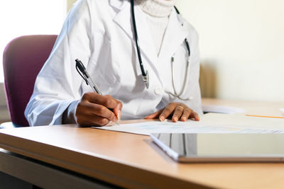 Crop unrecognizable black female physician with stethoscope writing information on paper sheet while preparing medical report at table in office of modern clinic