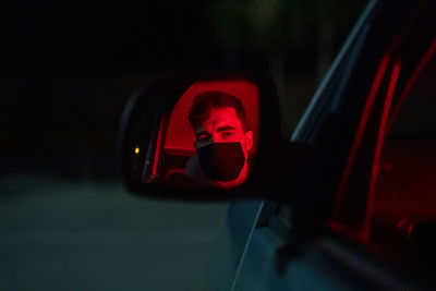 Anonymous serious male driver in face mask sitting in modern car with red neon light parked on street at night and looking in side mirror during pandemic