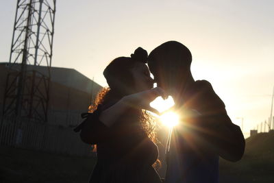 Couple making heart shape while kissing against bright sun during sunset