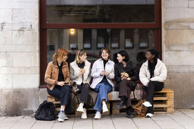 Happy female friends enjoying while sitting on pallets at sidewalk in city