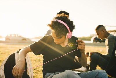 Teenage boy listening music on headphones while sitting with friend during sunny day