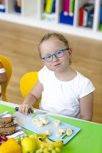 Portrait of cute girl holding knife while eating banana on table at school
