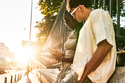 Low angle of diverse couple leaning on park fence and browsing cellphone together while spending time on city street at sunset