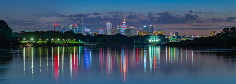 Illuminated buildings by lake against sky at night