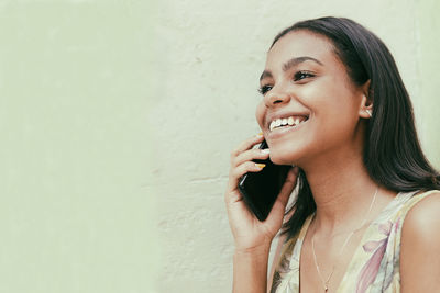 Smiling young woman looking away and using phone while standing against wall