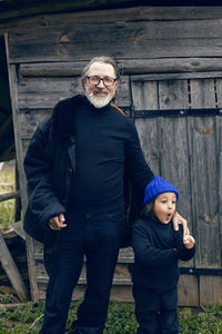 Grandson in a blue hat and boots with a grandfather in a sheepskin coat stand at a wooden house shed