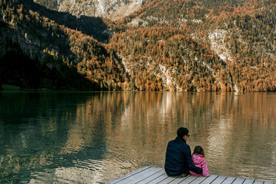 Rear view of father and daughter sitting by lake