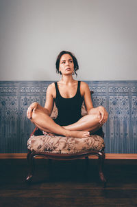 Young woman with legs crossed sitting on armchair against wall at home