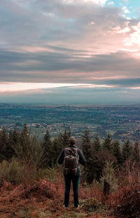 Rear view of man with backpack standing on mountain against cloudy sky during sunset