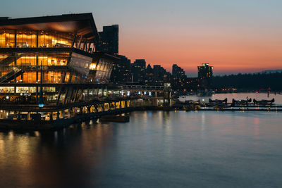 Illuminated buildings by river against sky at sunset