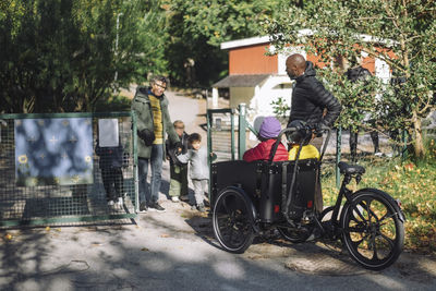 Female teacher standing at gate while welcoming children sitting in cargo bike at kindergarten