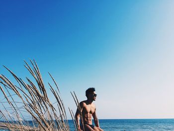 Shirtless young man sitting at beach against clear sky during sunny day