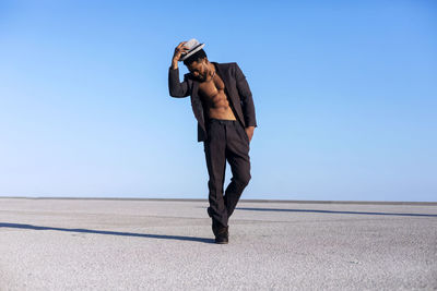 Full length of handsome man wearing hat standing on landscape against clear sky