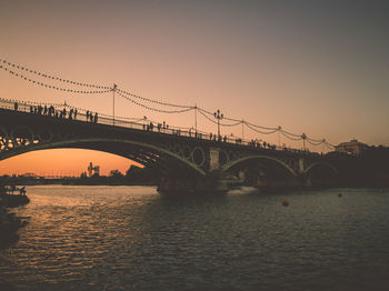 People on arch bridge over river against sky during sunset