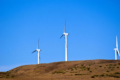 Low angle view of wind turbines on hill against clear blue sky