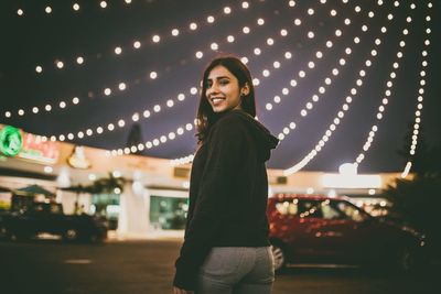 Portrait of smiling young woman standing against illuminated city at night