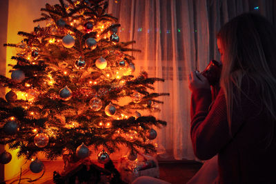 Woman drinking coffee by illuminated christmas tree