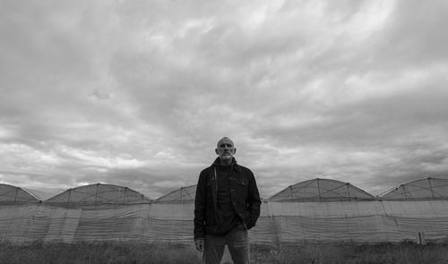 Adult man standing in front of plastic greenhouses against cloudy sky in almeria, spain