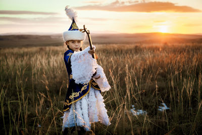 Young woman with arms raised standing on field against sky during sunset