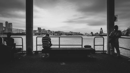 Man sitting on railing by river in city against sky