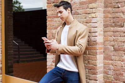 Young man using phone while standing by brick wall